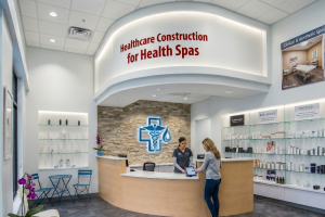 Reception area of a modern medical spa with a curved white desk, stone accent wall, and a glowing blue medical cross logo behind the counter; a staff member assists a visitor at the desk.