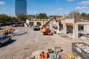 Construction site with workers in safety gear on scaffolding and an orange boom lift building a concrete structure; a tall blue glass building in the background.