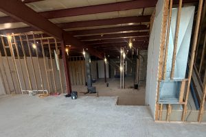 Unfinished interior under construction with exposed wooden studs, support columns, and hanging work lights.