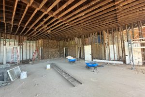 Interior of a building under renovation with exposed wooden studs, ceiling joists, and scattered ladders and wheelbarrows.