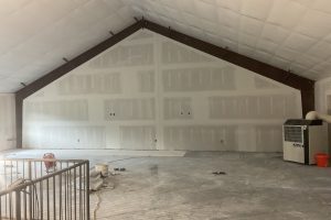 Unfinished interior of a large room with a peaked ceiling, white drywall with taped seams, brown beams, and construction debris on the floor; an air conditioner unit is on the right.