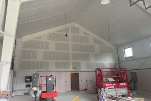 Interior of a large workshop under construction: drywall panels on the far wall, a red elevated work platform, and various tools scattered around.
