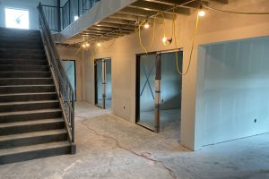 Under‑construction interior: concrete staircase on the left, exposed wiring and hanging lights, and multiple open doorways with blue painter’s tape X marks on the walls.