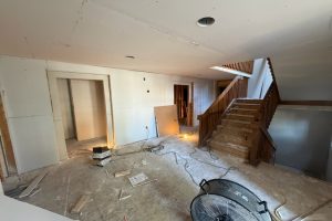 Interior under renovation with drywall panels, exposed framing, and a wooden staircase under construction. Debris and a large fan on the floor.