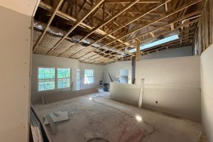Unfinished interior during renovation: exposed wooden ceiling framing and ductwork, partially installed drywall, construction debris on the concrete floor, two windows letting in light, and a temporary wall panel.