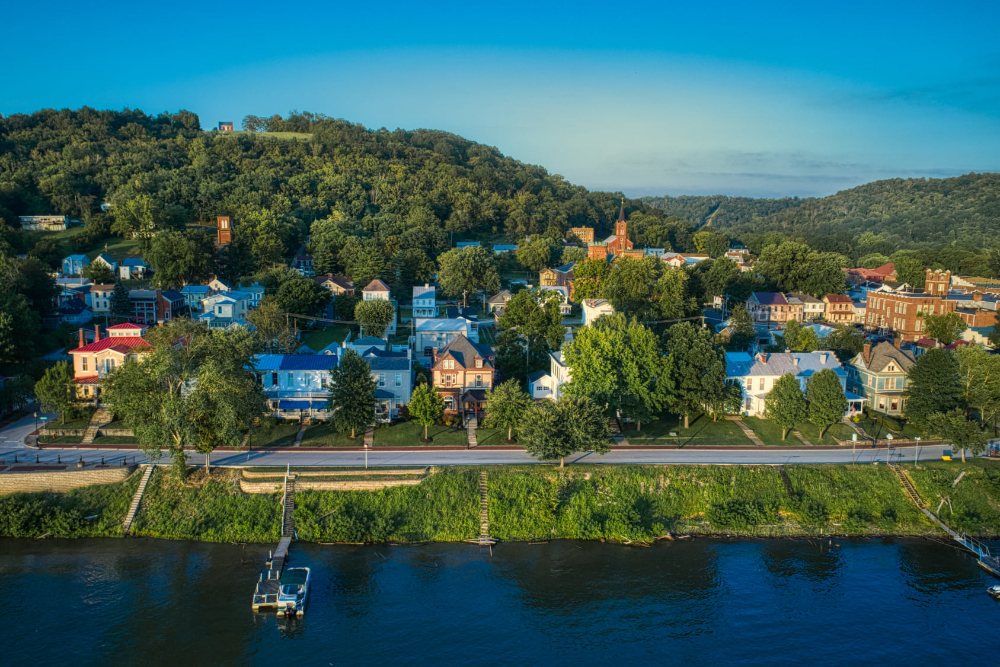Aerial view of a riverfront town with colorful houses, tree-lined streets, and green hills in the background by the water.