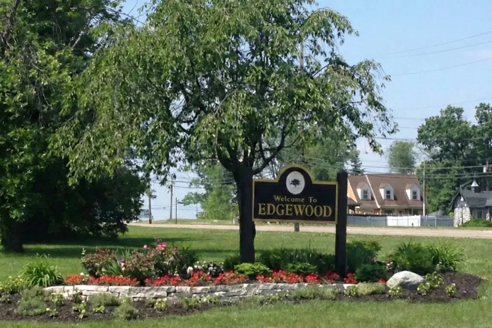 Welcome sign for Edgewood, black panel with gold lettering, set in a landscaped flower bed with trees nearby.