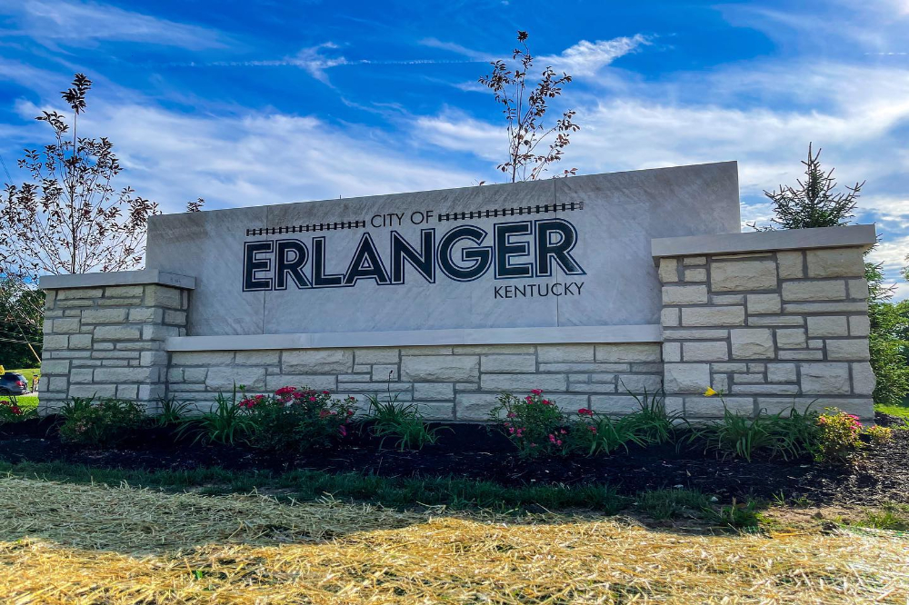Large stone roadside sign reading 'City of Erlanger Kentucky' with a landscaped flower bed in front and a bright blue sky behind it
