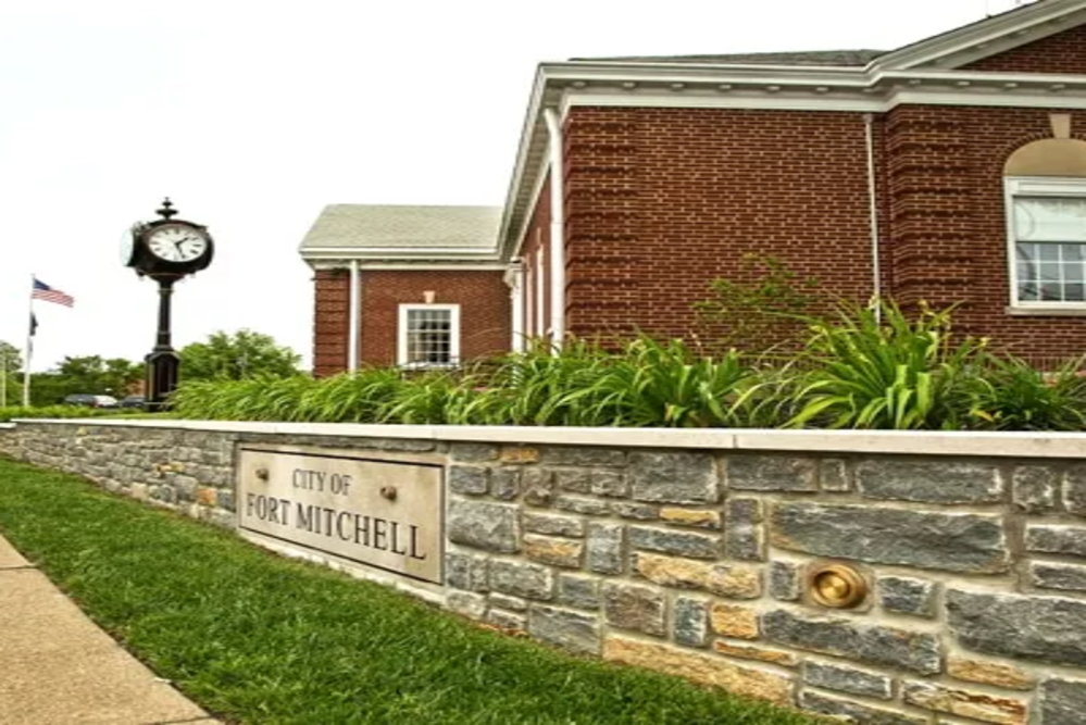 Large stone roadside sign reading 'City of Erlanger Kentucky' with a landscaped flower bed in front and a bright blue sky behind it