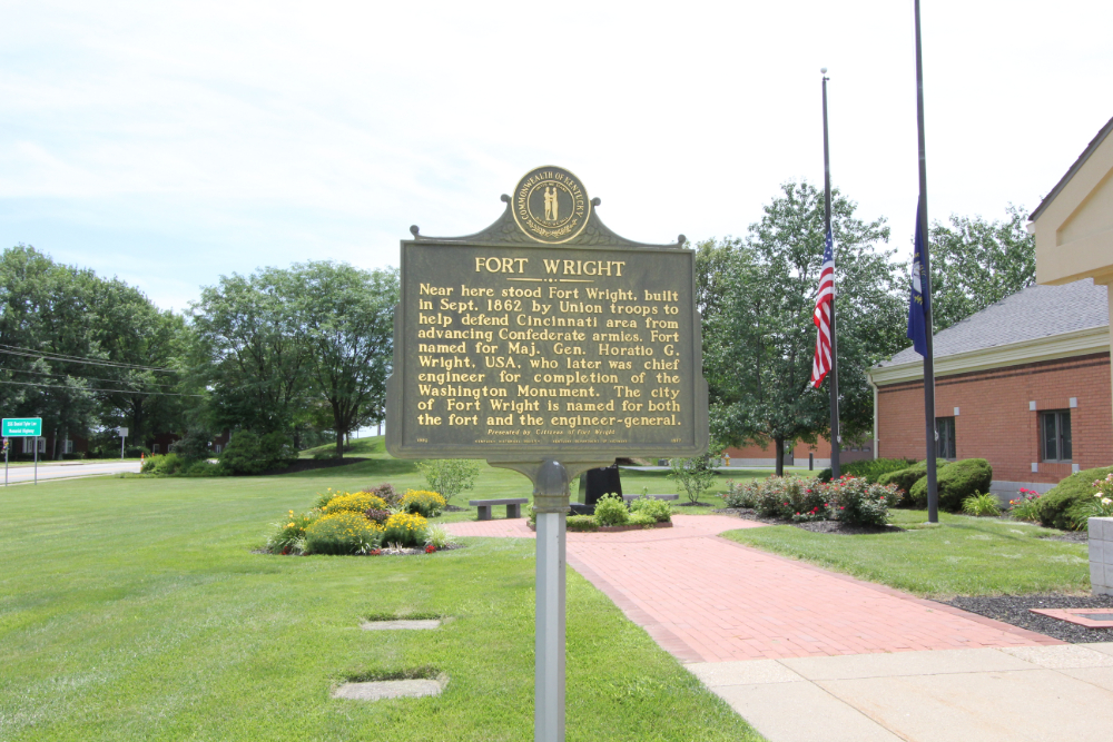 Large stone roadside sign reading 'City of Erlanger Kentucky' with a landscaped flower bed in front and a bright blue sky behind it