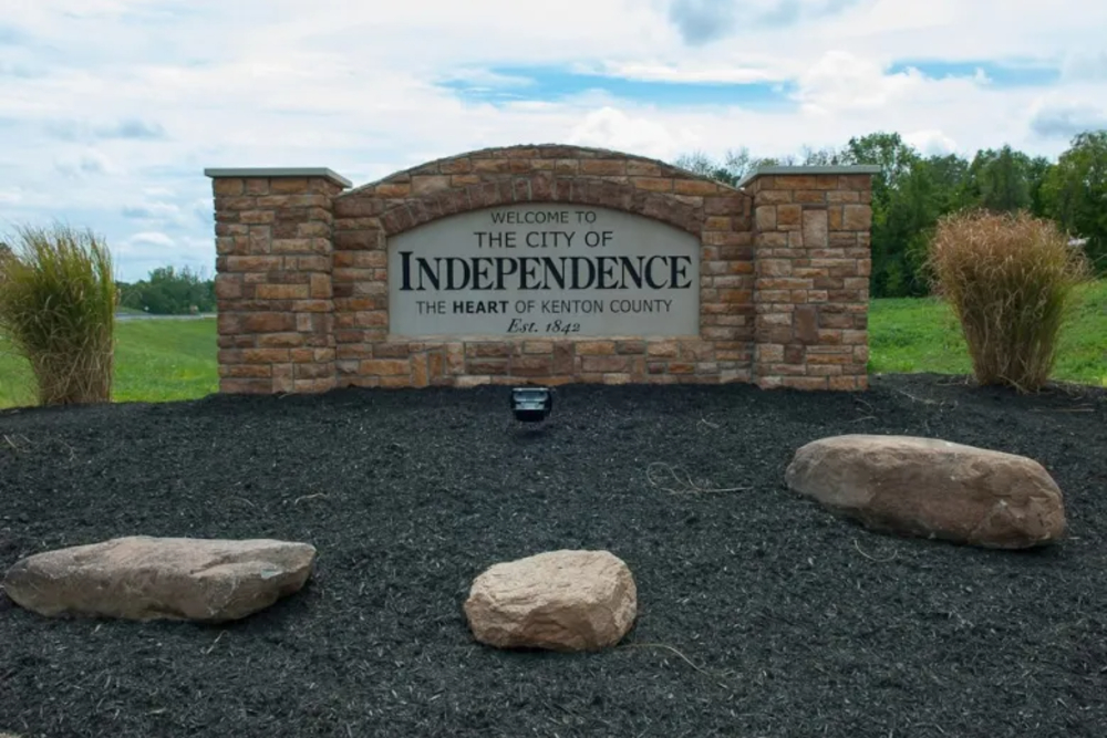 Large stone roadside sign reading 'City of Erlanger Kentucky' with a landscaped flower bed in front and a bright blue sky behind it